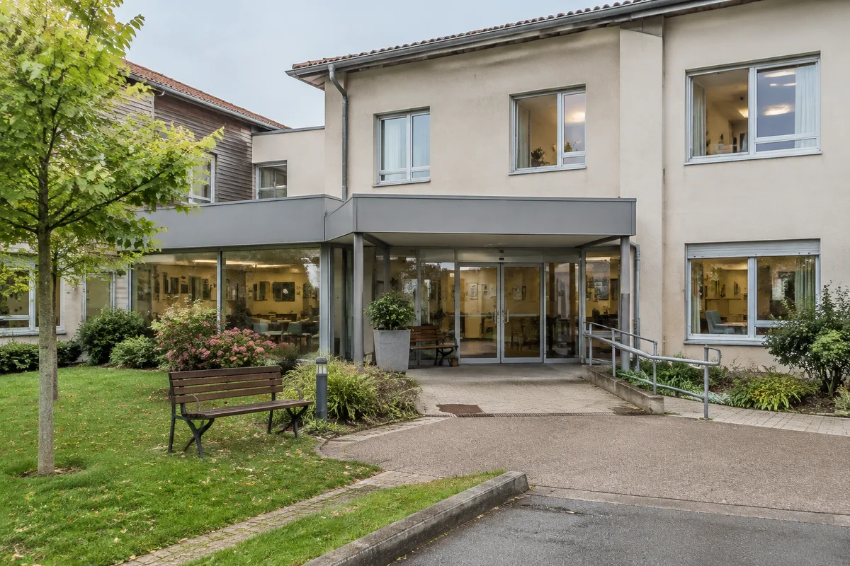 Façade réaliste d’un EHPAD médicalisé avec entrée, jardin et espaces communs